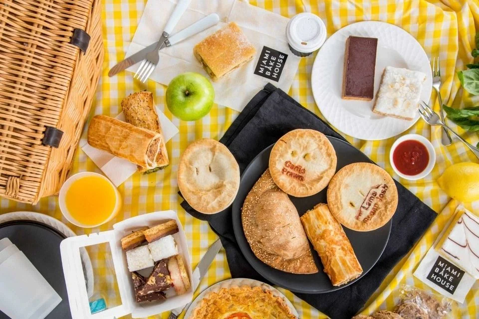 An overhead shot of a picnic on a yellow checkered cloth, featuring various pies, pastries, quiche, dessert bars, an apple, bread rolls, sauce, and drinks arranged neatly.