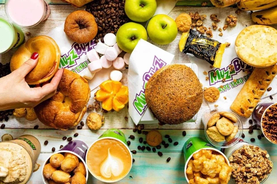 A colorful spread of breakfast foods, including bagels, muffins, cookies, apples, coffee drinks, granola bars, marshmallows, and pastries, with a hand reaching for a bagel. Muzz Buzz branding is visible on several items.