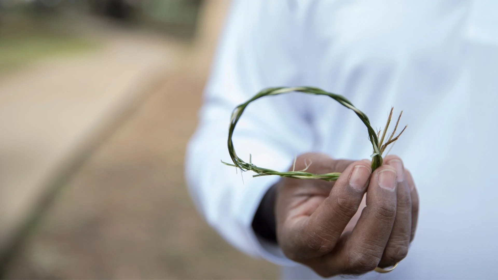 A person in a white shirt holds a twisted loop made of long green grass strands. The background is blurred, showing a pathway and natural surroundings.