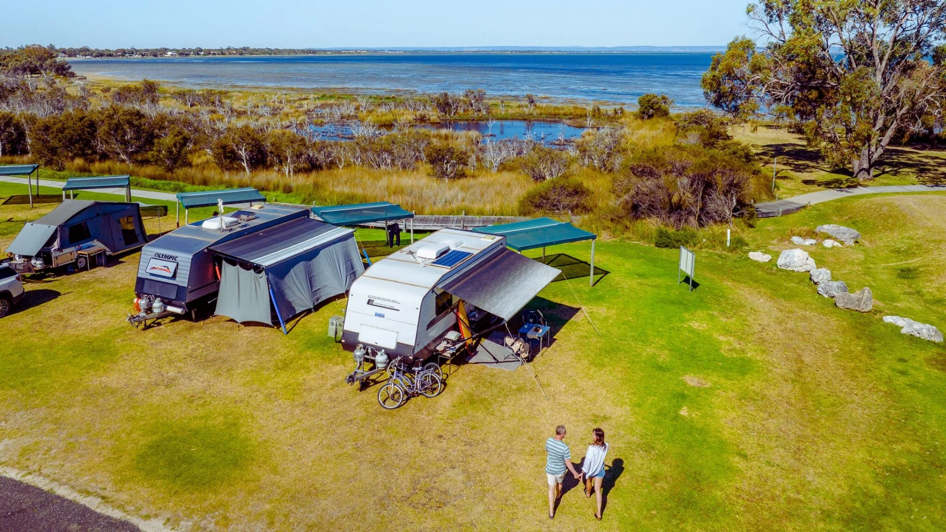Aerial view of a campsite by a lake. Two people walk towards three caravans with awnings. Bicycles are parked nearby. The area is grassy with scattered trees, and the lake is in the background.
