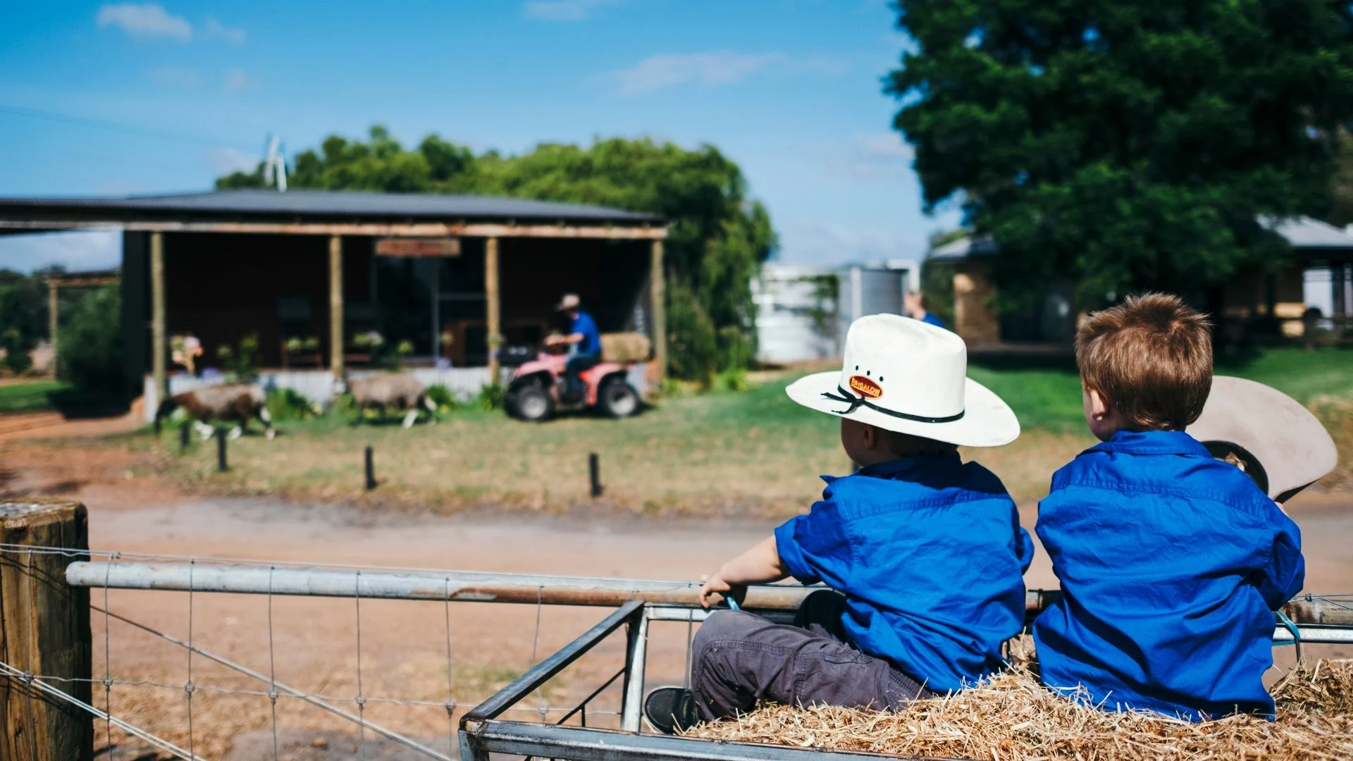 Two children in blue shirts and hats sit together on a hay bale in a farm setting. They watch a person on a vehicle near a barn. The background has trees and a clear sky.
