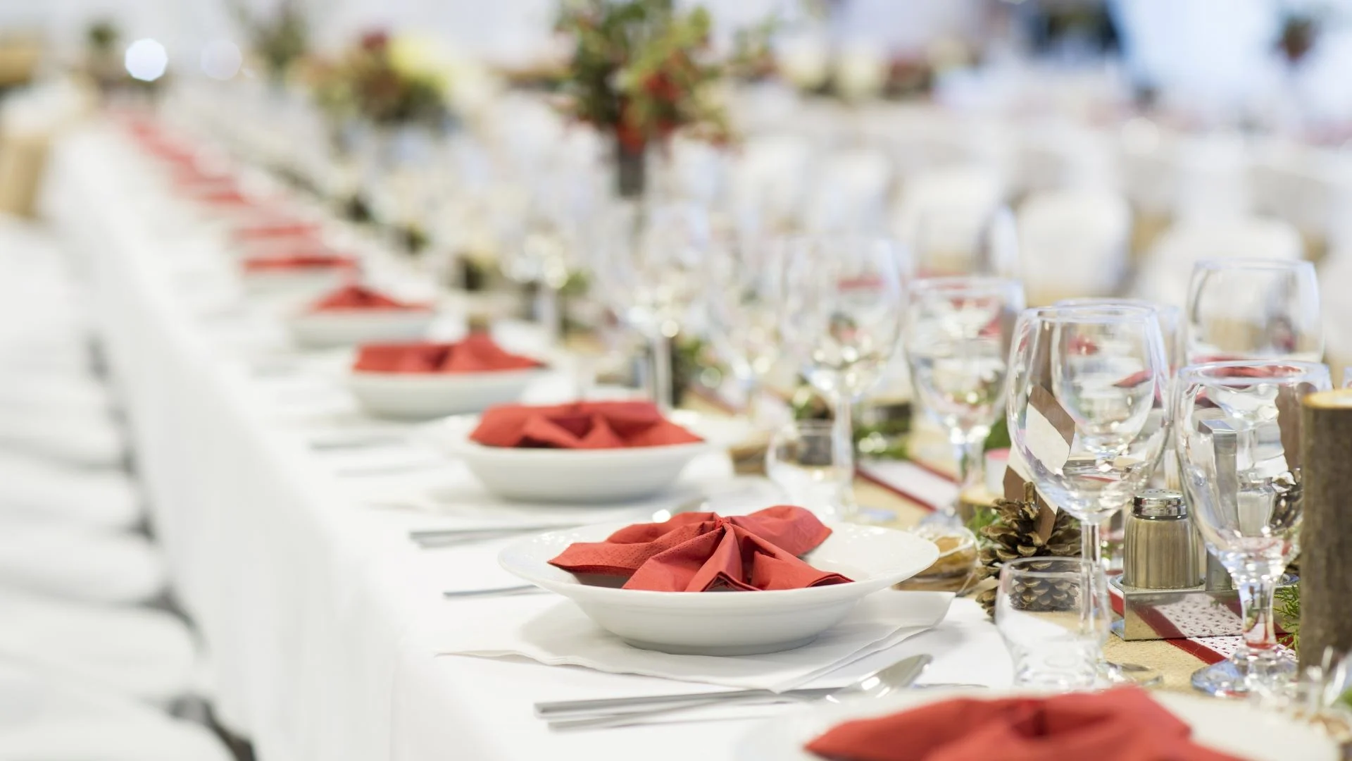 A long banquet table set for an event, featuring white tablecloths, neatly folded red napkins in white plates, wine glasses, cutlery, and floral centerpieces. The table extends into the blurred background.