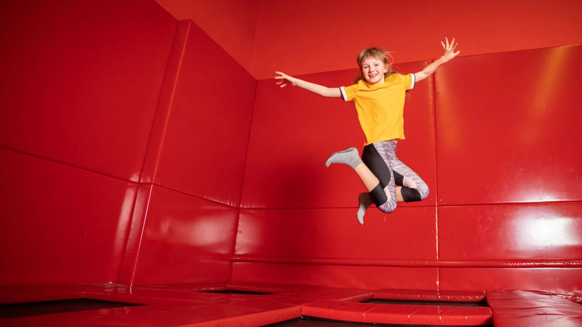 Child in a yellow shirt and blue socks joyfully jumps on a trampoline inside a bright red indoor space, with arms and legs outstretched.