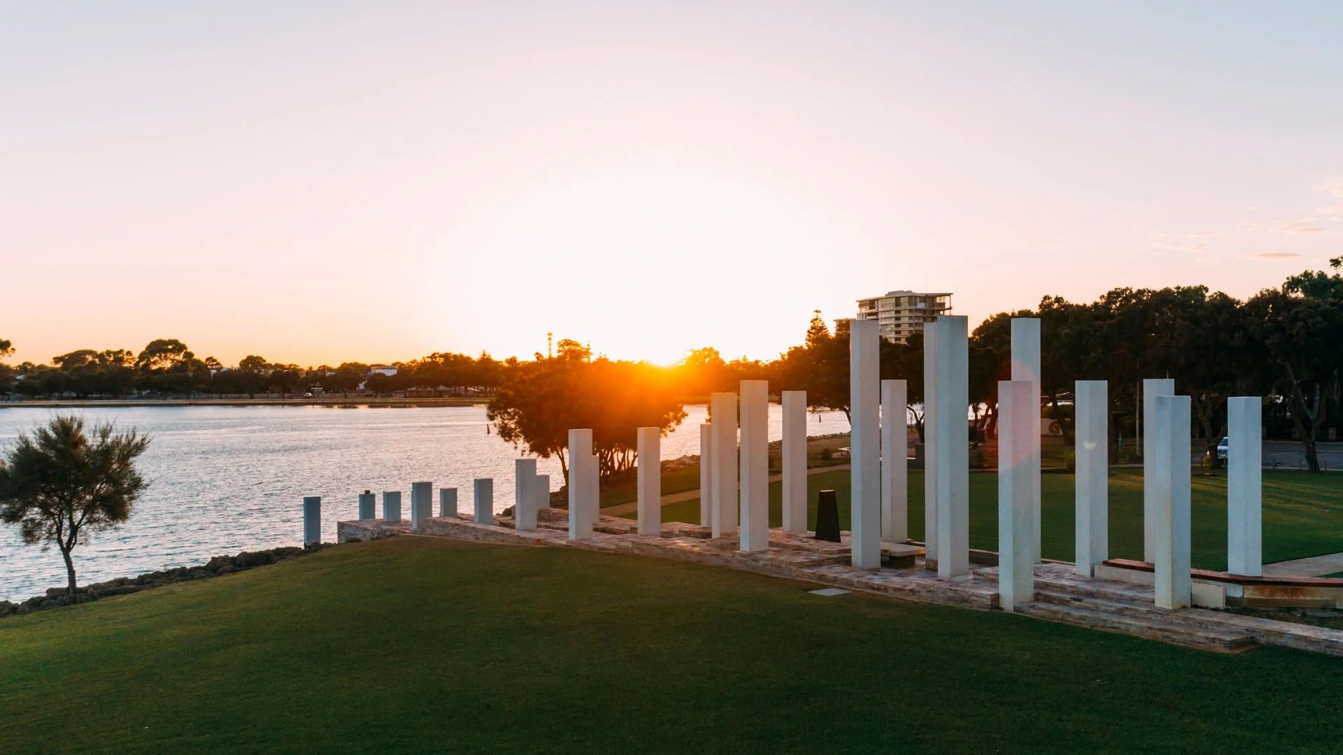 A scenic view of a waterfront park at sunset, featuring tall, narrow stone pillars arranged in a row on a grassy area. The sun is setting on the horizon, casting a warm glow over the water and trees in the background.