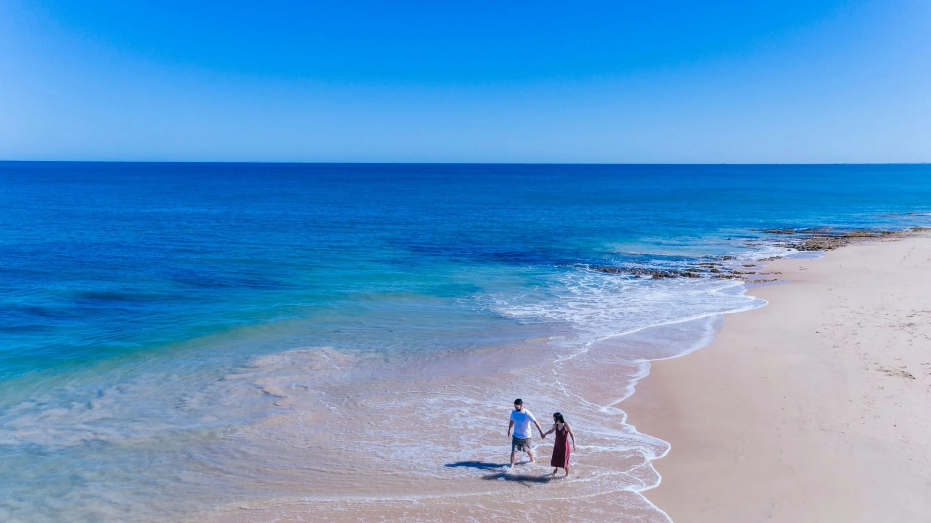 A couple walks hand in hand along the shoreline of a sandy beach, with clear blue ocean waves gently washing up beside them under a bright blue sky.