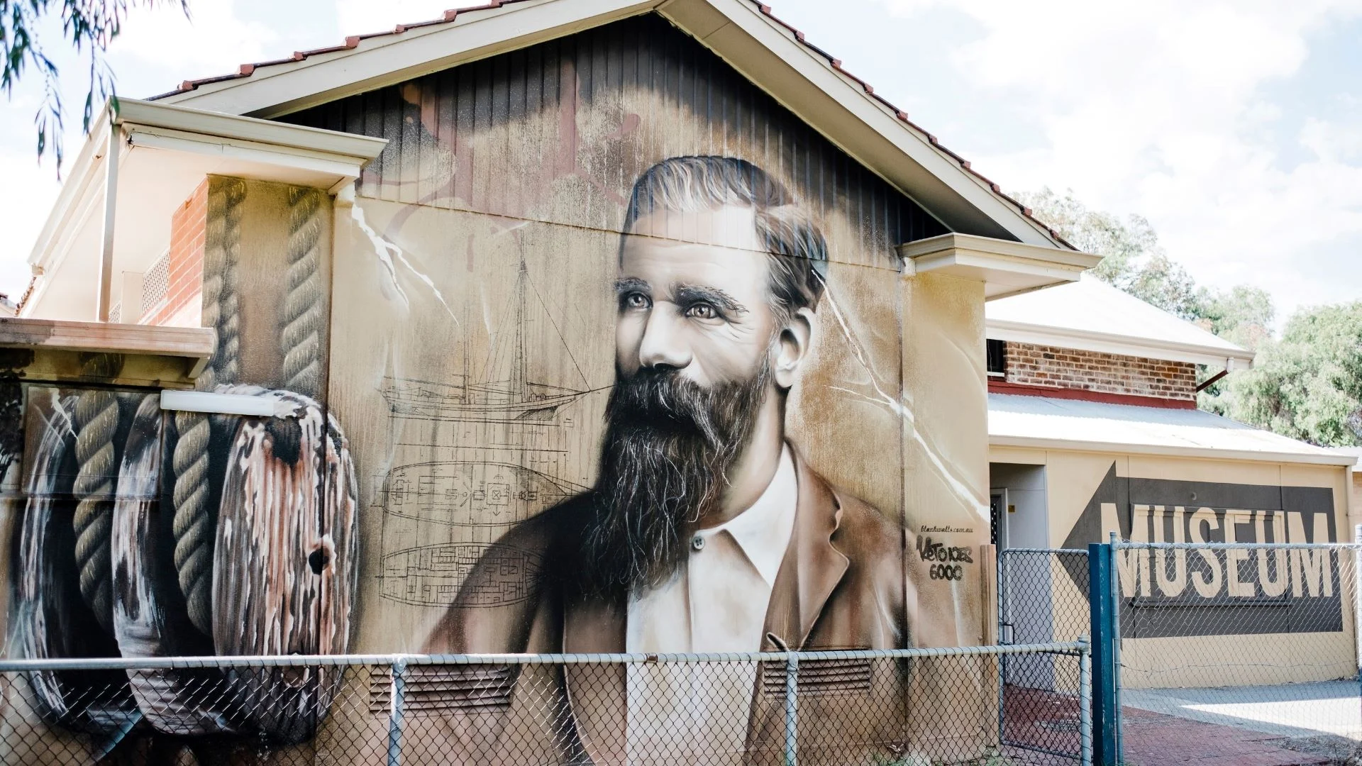 Mural of a bearded man with a detailed sketch behind him on the side of a house in an outdoor museum setting. The artwork blends realistic portraiture with architectural drawings. A partially visible MUSEUM sign is in the background.