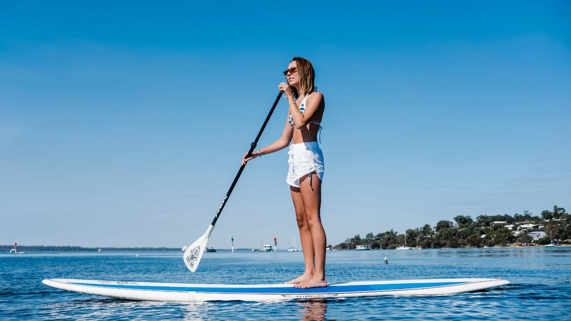 A woman wearing sunglasses, a white top, and white shorts stands on a paddleboard, holding a paddle on calm blue water with a clear sky and distant shoreline in the background.