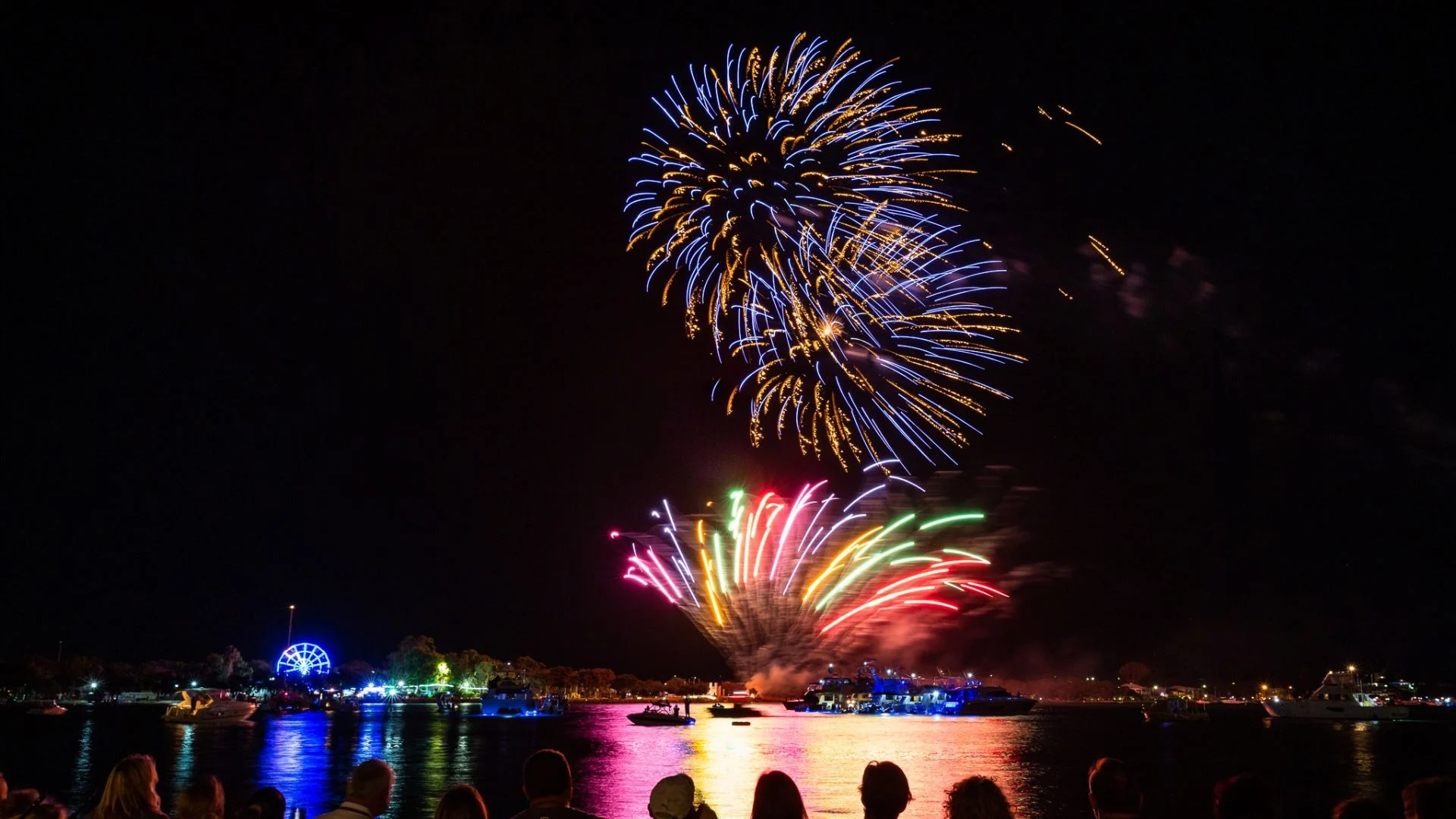 Colourful fireworks burst in the night sky over a waterfront, reflecting on the water, as silhouetted people watch from the shore. A lit ferris wheel and boats are visible in the background.