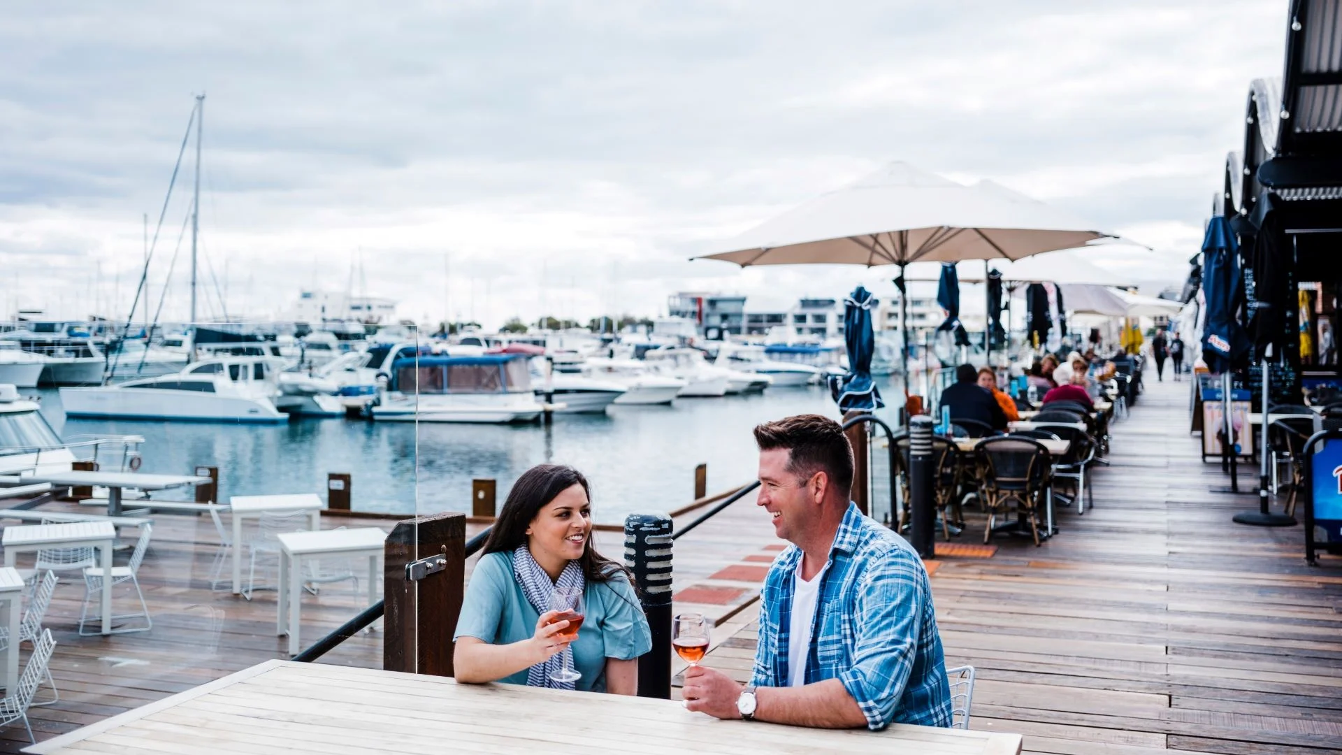 A man and woman sit at an outdoor table by a marina, smiling and holding drinks. Boats are docked in the water and people are dining along a wooden boardwalk under umbrellas on a cloudy day.