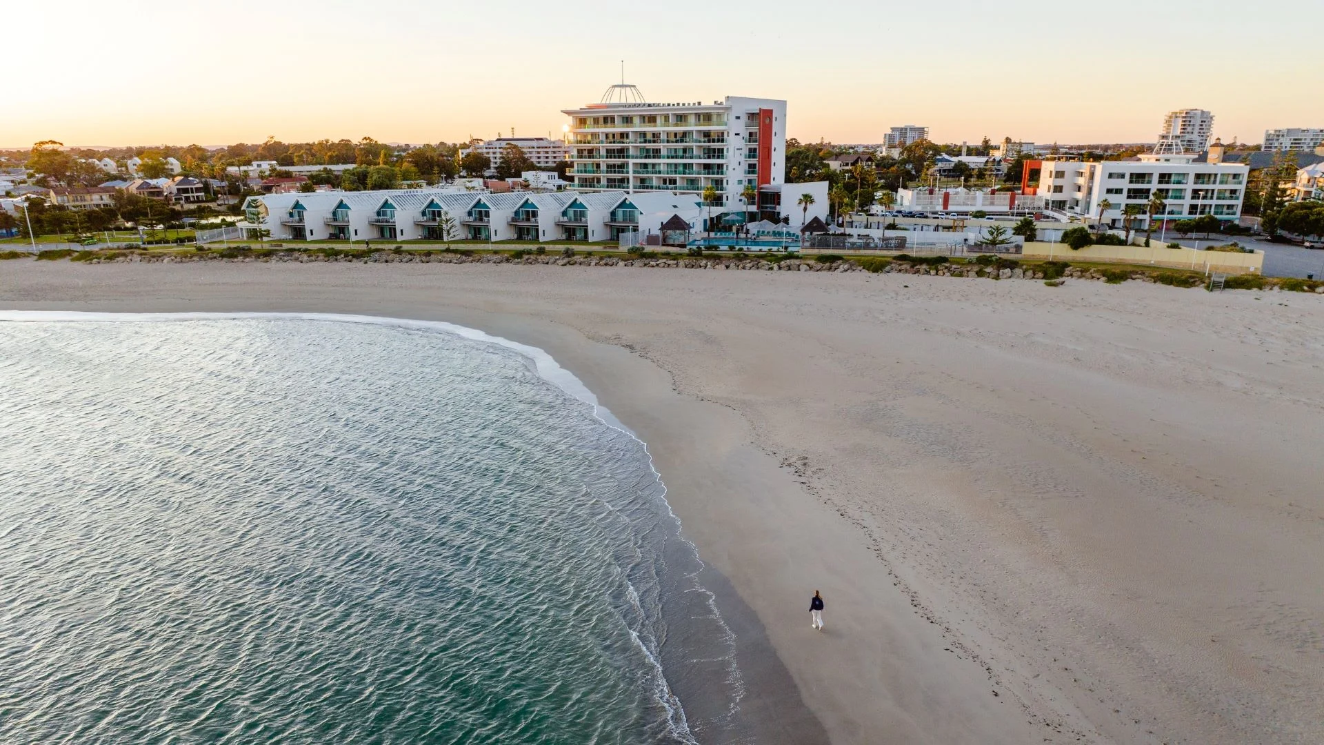 A wide, sandy beach with gentle waves, bordered by modern beachfront buildings; a single person walks along the shore in the early morning or late evening light.