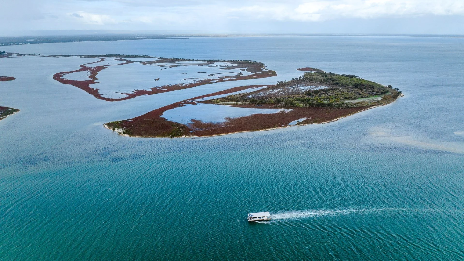 A small white houseboat moves across turquoise water near a sparsely vegetated island with red and green patches, surrounded by more water and distant land under a cloudy sky.