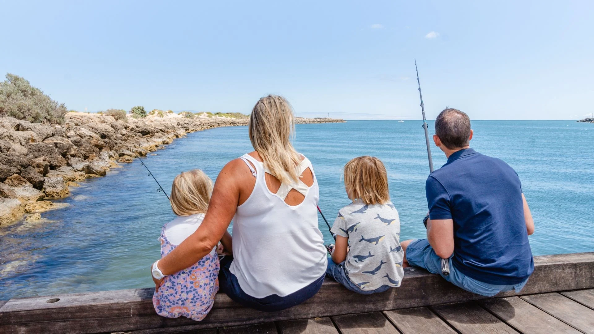 A family of four sits on a wooden pier, fishing and enjoying a sunny day by the water. The mother has her arm around a young girl, while the father and another child sit nearby. The sea is calm with rocky shores in the background.