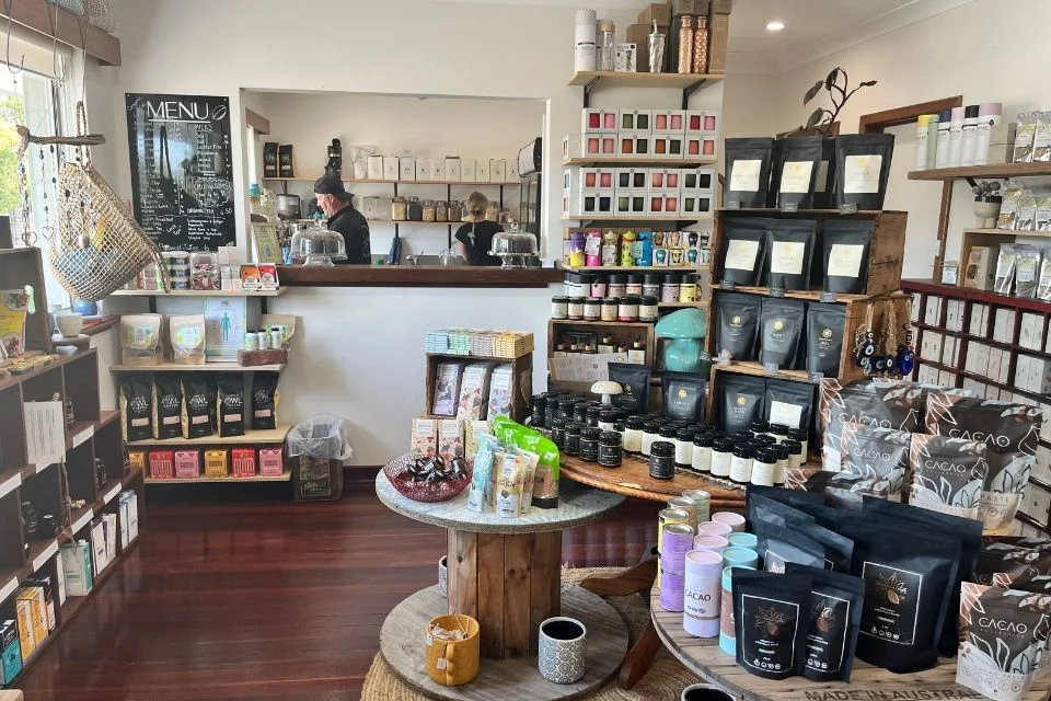 A cosy shop with shelves filled with tea bags, candles, and various products. A chalkboard menu is visible. A person is working behind the counter. The wooden floor and bright sunlight give the space a warm atmosphere.