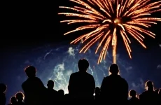 A group of people silhouetted against the night sky watch bright orange fireworks bursting above them.
