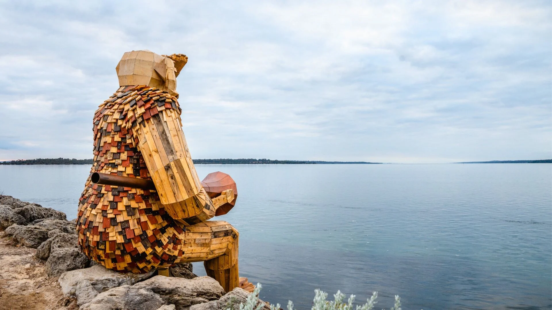 Large wooden sculpture of a seated figure with a patchwork surface, overlooking a body of water. The figure appears to be gazing into the distance with a peaceful backdrop of a cloudy sky and distant shoreline.