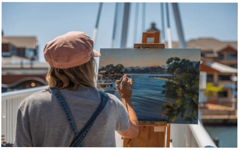 A person wearing a pink cap and gray shirt paints a scenic lakeside view on a canvas set on an easel outdoors, with buildings and water visible in the background.