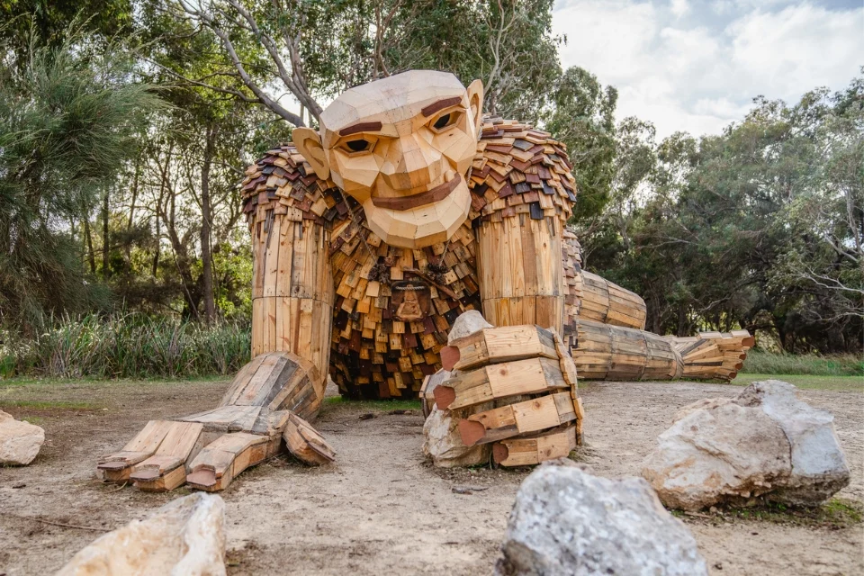 A large wooden sculpture of a troll-like figure crouches on the ground, surrounded by trees and rocks. Made from variously sized wooden planks, the sculpture features a detailed face and limb structures. The sky is partly cloudy.