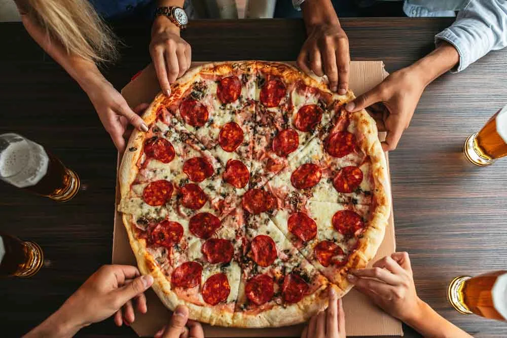 A group of people reaching for slices of a large pepperoni pizza on a wooden table. Glasses of beer are placed around the pizza box, suggesting a casual gathering or meal. The photo is taken from an overhead perspective.