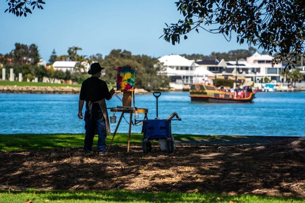 A person in a hat paints on an easel by a riverbank, with a colourful sailboat passing on the water. Houses are visible in the background, and trees frame the scene. The ground is covered with grass and scattered leaves.