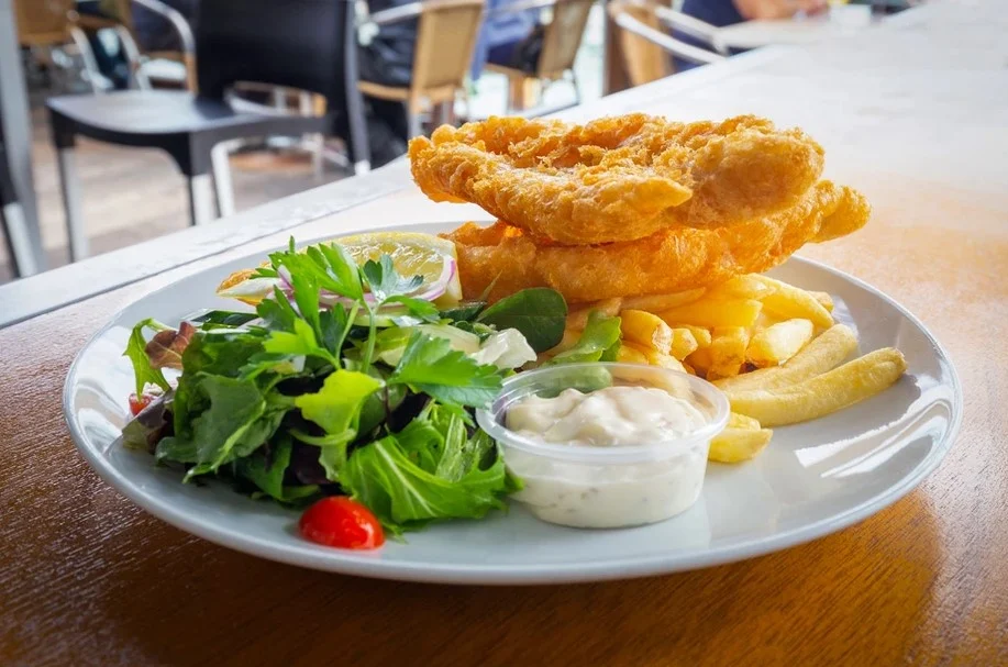 A plate of fish and chips with a side of mixed salad and a small container of tartar sauce. The fish is golden brown and crispy, served with lemon wedge and thick-cut fries. The plate is set on a wooden table in an outdoor dining area.