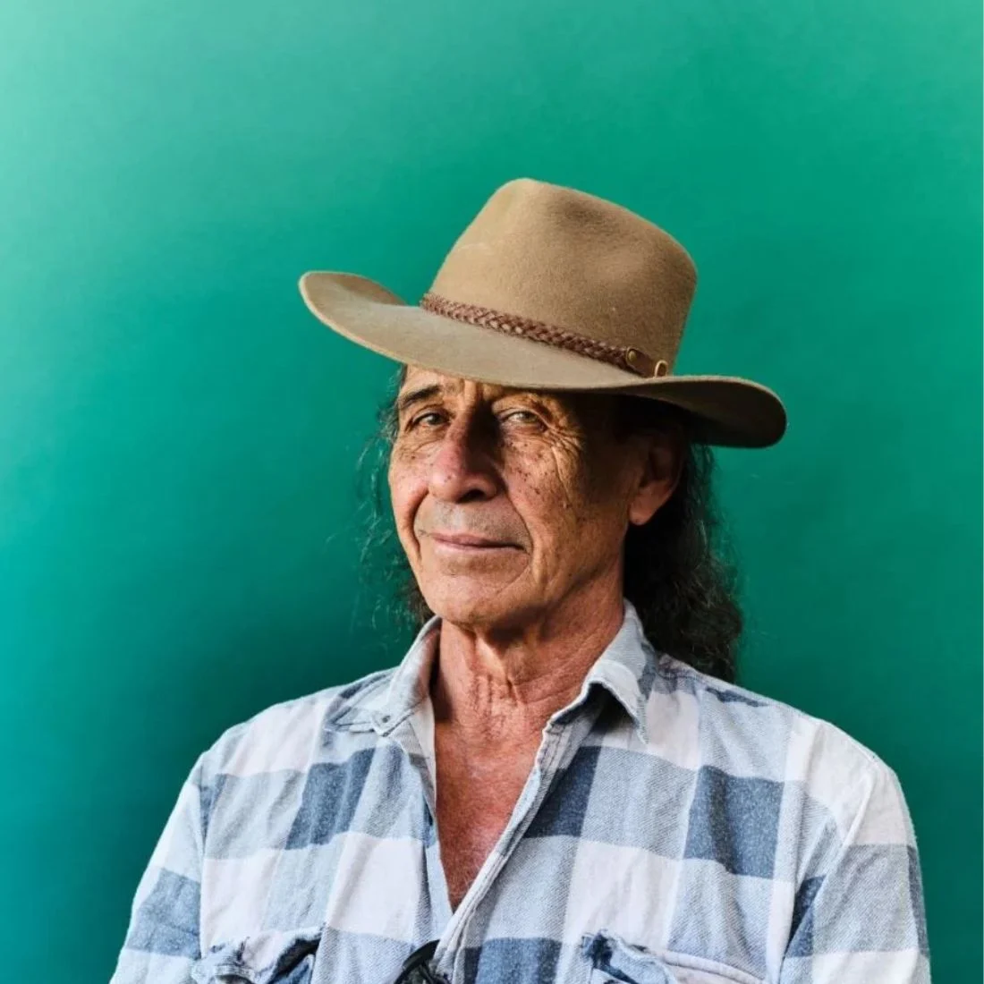An older man with long hair wears a tan hat and a blue-and-white plaid shirt, standing in front of a plain teal background. He has a calm expression and looks slightly to the side.