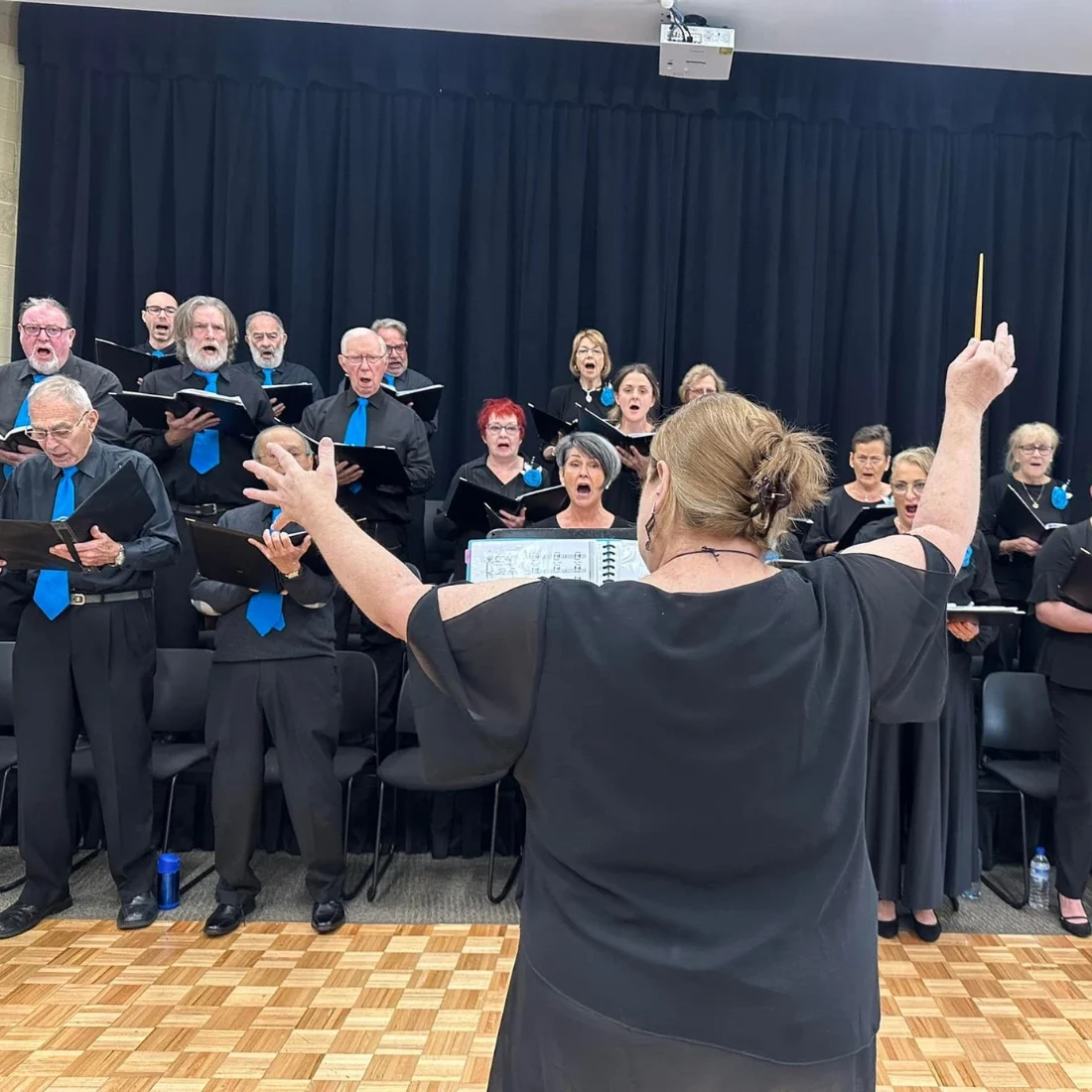 A choir of men and women dressed in black, some with blue ties or pins, sings from sheet music as a conductor stands facing them with raised arms, directing the performance on a wooden parquet floor.