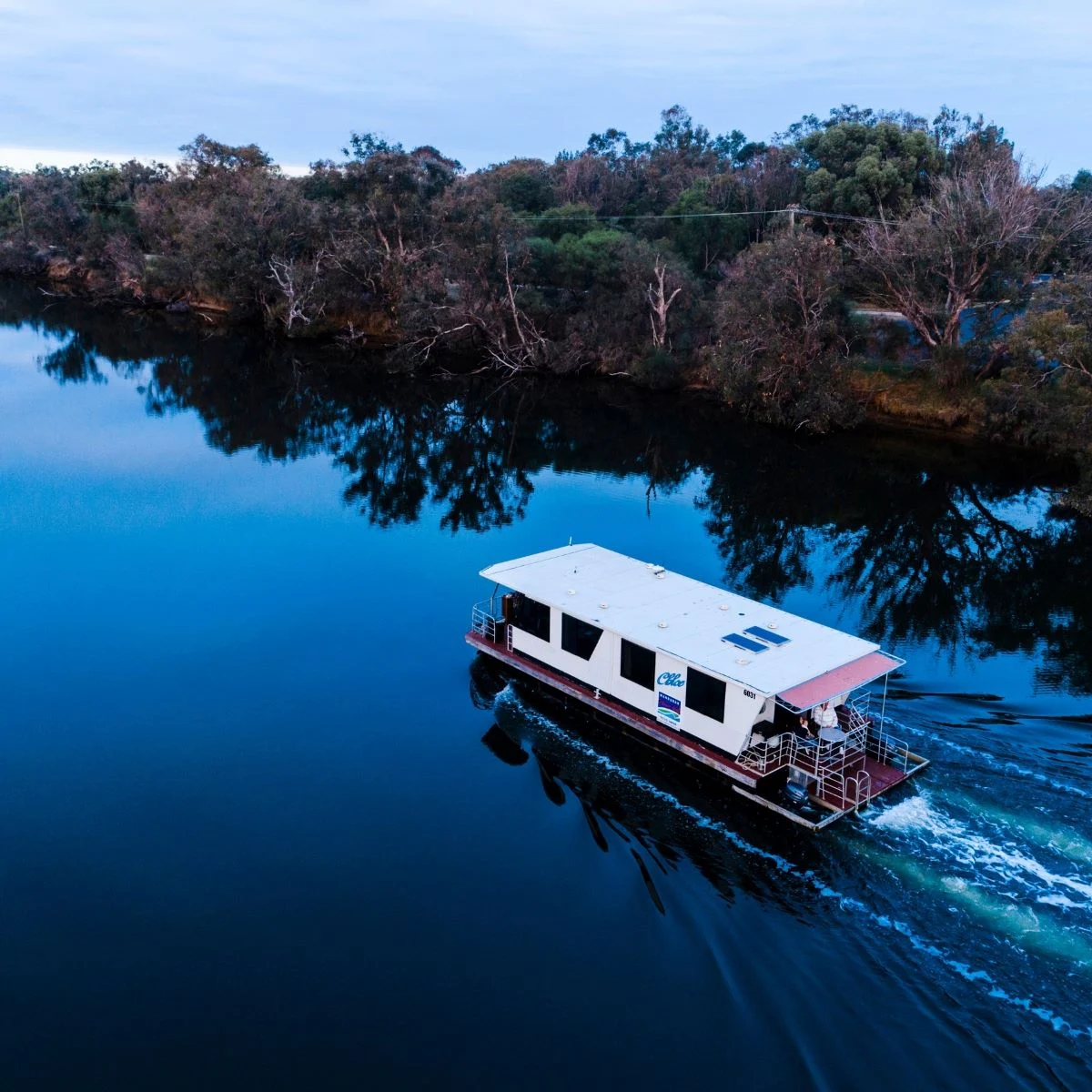 A white and red houseboat travels along a calm, reflective river bordered by trees, under a cloudy sky.