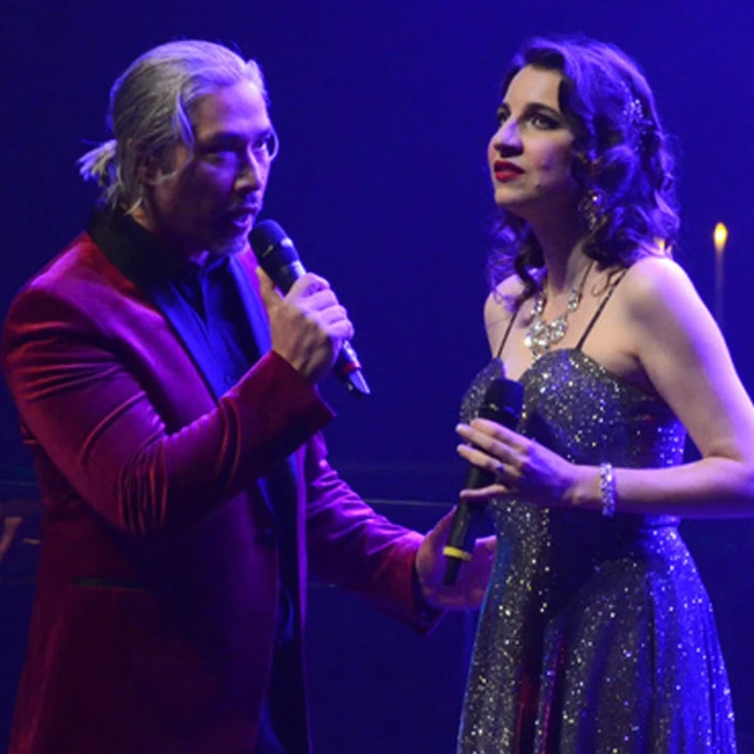 A man in a red velvet suit jacket and a woman in a sparkly dress sing into microphones on a dimly lit stage, with purple lighting and candles in the background.