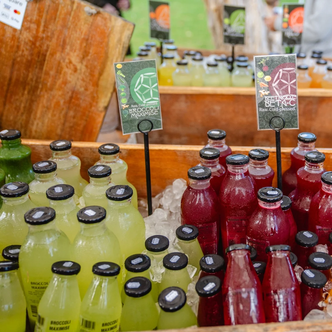 Bottles of green and red cold-pressed juice are displayed on ice at a market stand, each labeled with a sign showing the juices ingredients. The green juice is labeled Broccoli Magnifique and the red juice Redroot Beta-C.