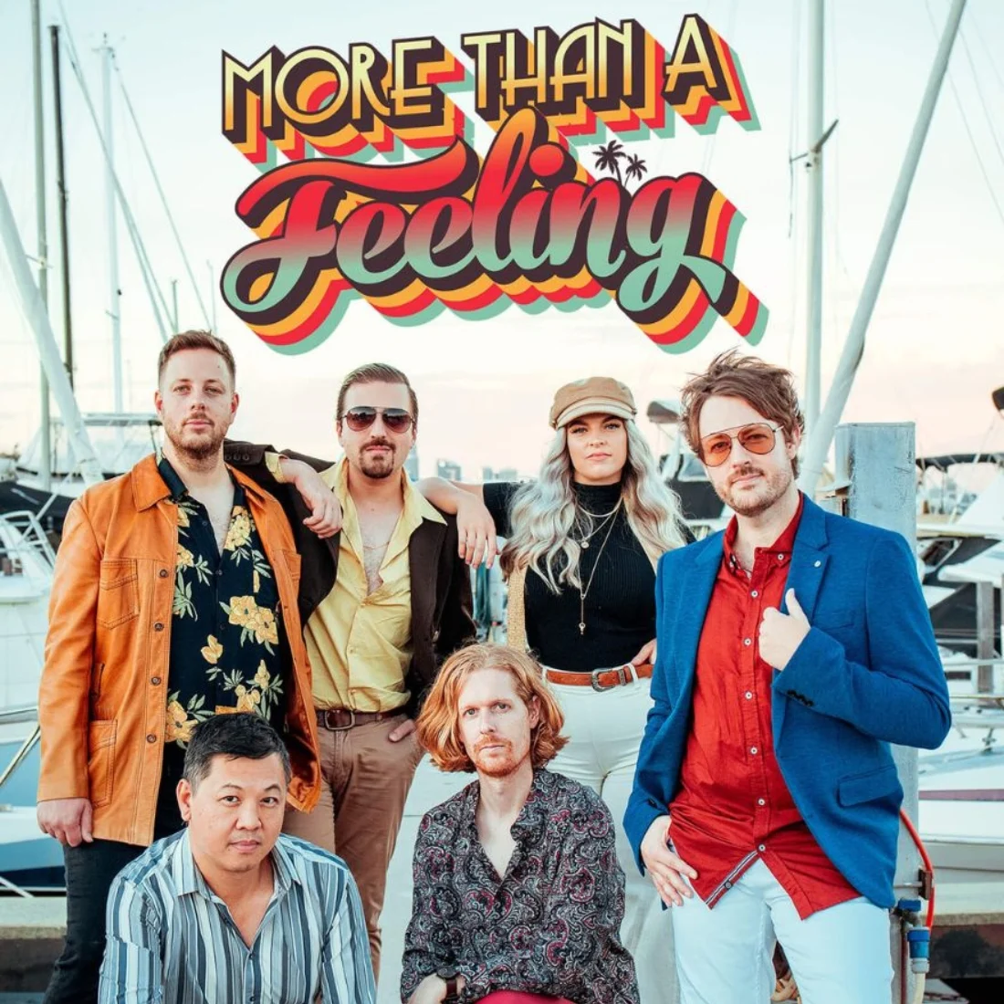 Six people pose by a marina with sailboats, dressed in retro 1970s-style clothing. Above them, colorful, bold text reads “More Than A Feeling.” The group stands confidently, with a sunset sky in the background.