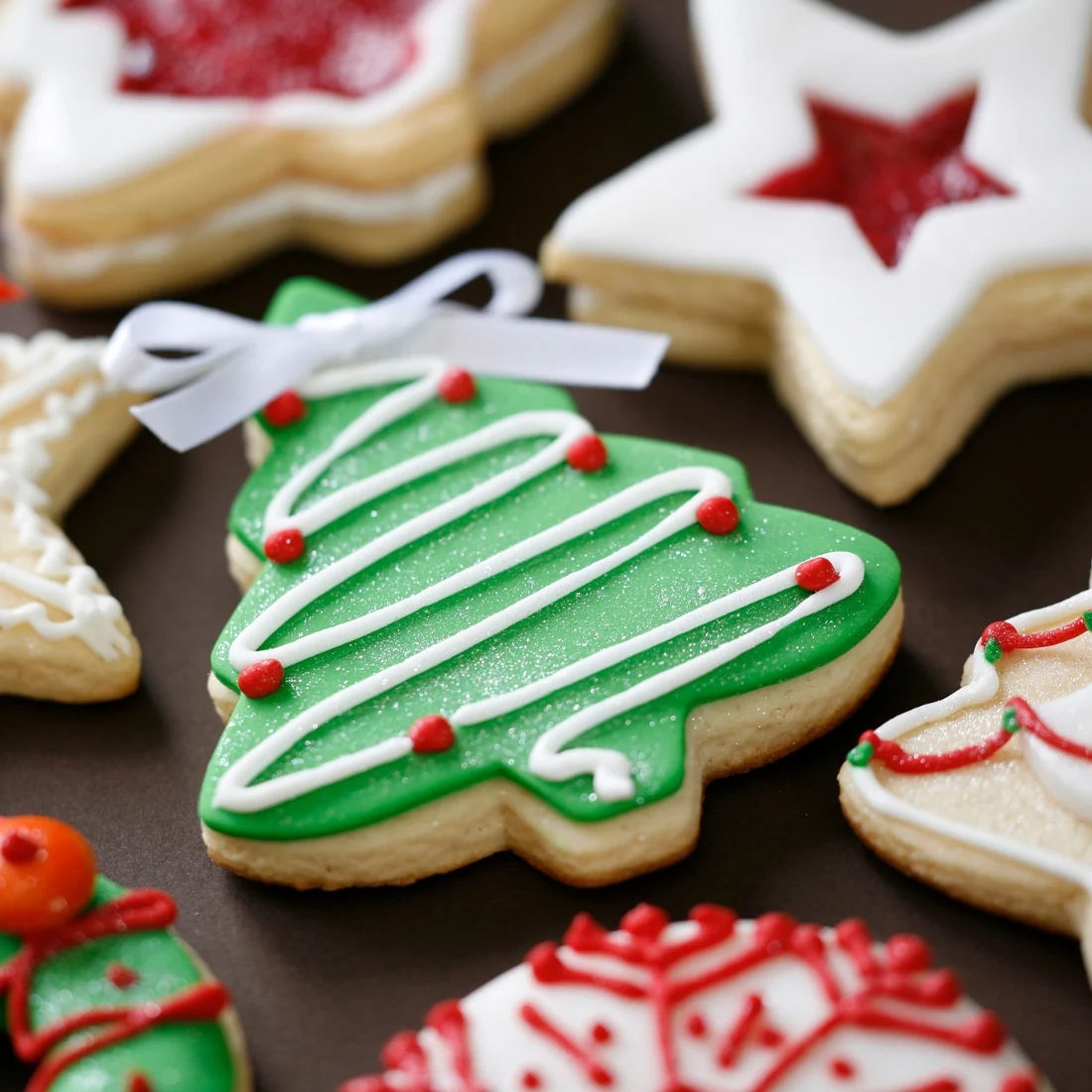 Assorted decorated Christmas cookies, including a green Christmas tree cookie with white icing, red dots, and a small white bow, surrounded by star-shaped and round cookies with festive icing.
