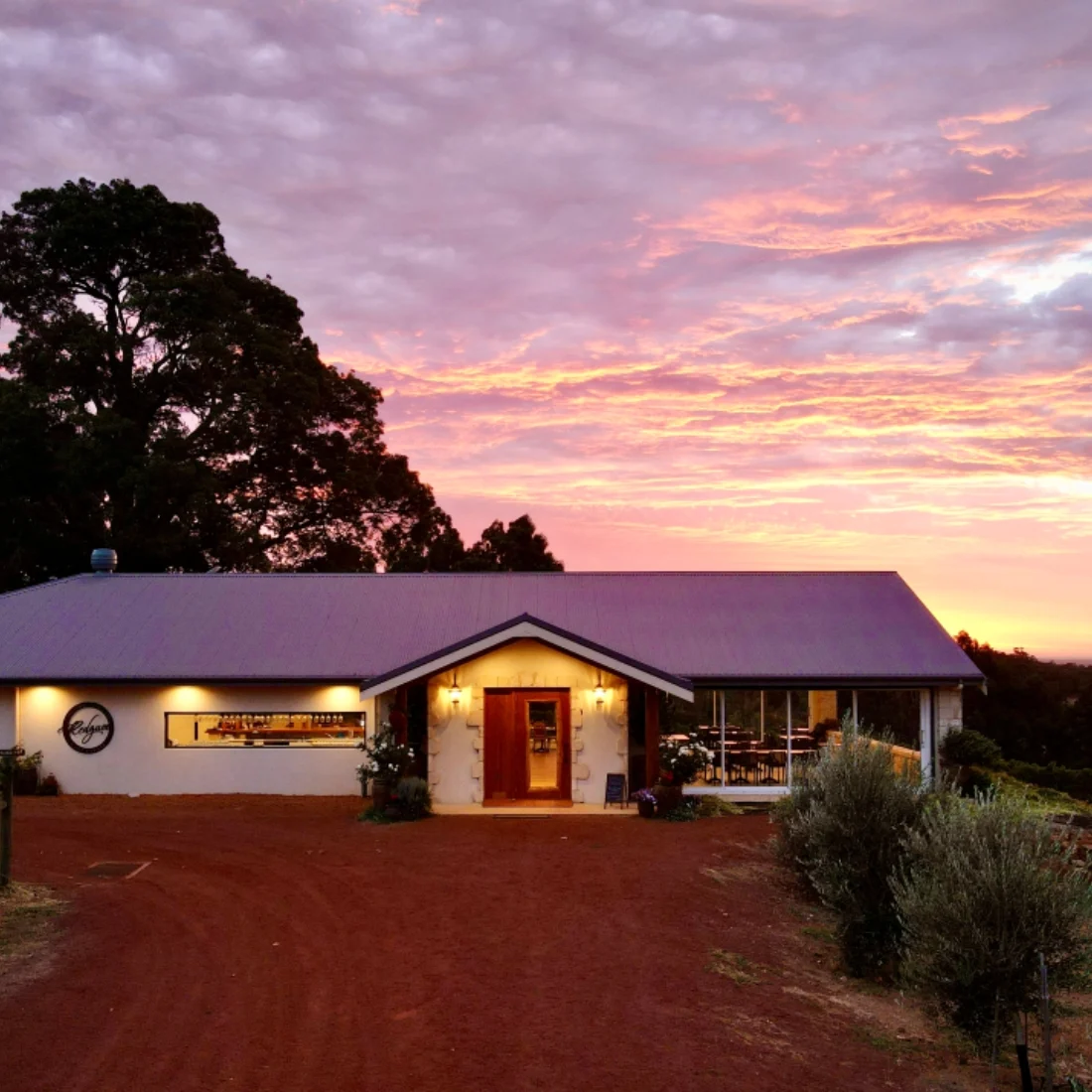 A rustic house with warm lights glows under a vibrant, colorful sunset sky. The building sits on red dirt with some plants and trees around it, creating a peaceful, rural atmosphere.