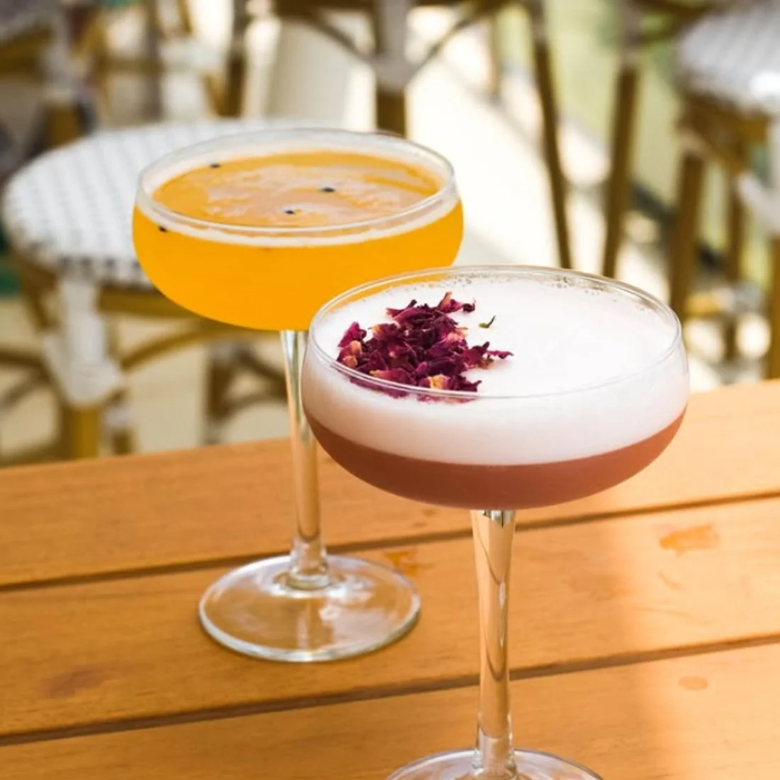Two elegant cocktails on a wooden table: one yellow-orange with small garnish, and one pink with a thick frothy white top adorned with dried flower petals. Blurred chairs are visible in the background.