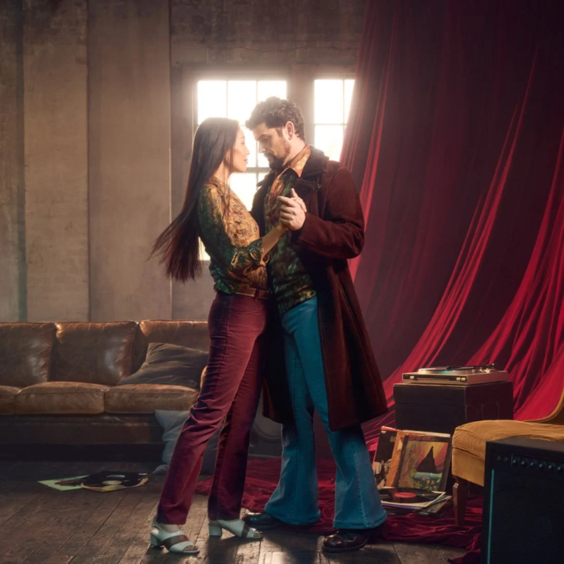 A couple dressed in retro 1970s-style clothing dances closely in a dimly lit room with vintage decor, including a brown sofa, vinyl records, a turntable, and red draped curtains.
