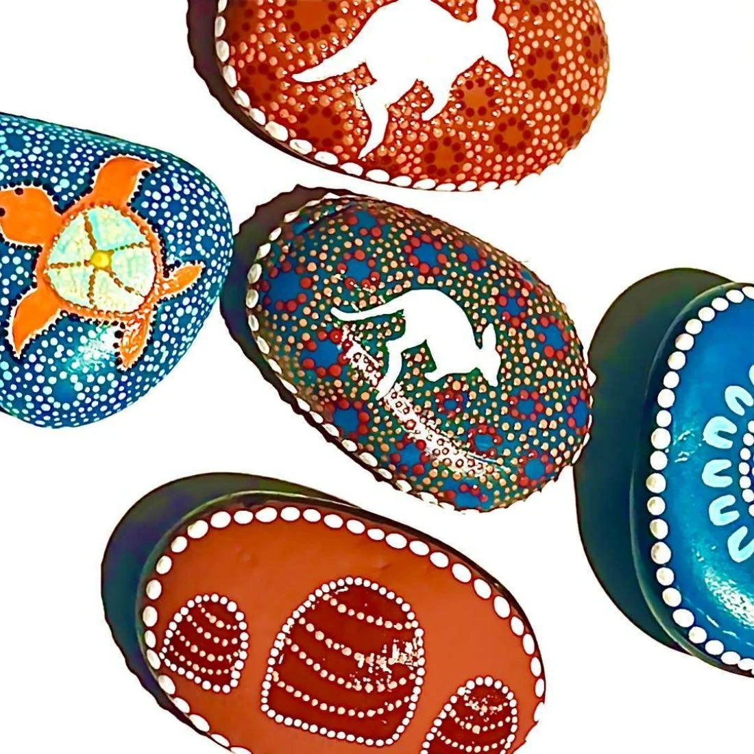 Colorful rocks painted with dot art patterns featuring animals such as a kangaroo, turtle, and a hive. The stones have intricate designs and vibrant colors on a white background.
