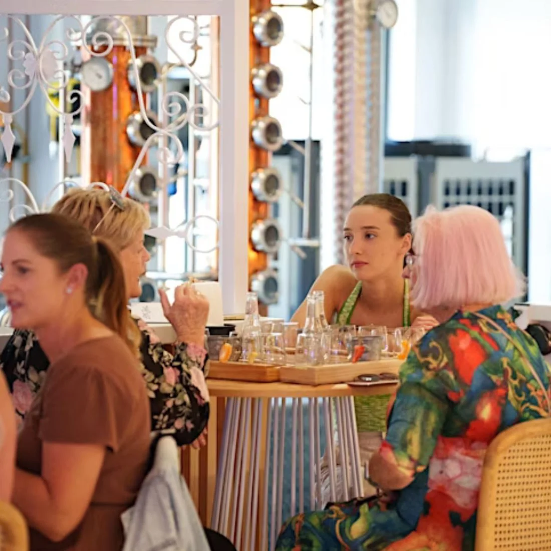 Four women sit around a table in a brightly lit restaurant, engaged in conversation. The table has glasses and tasting paddles, and the background shows copper distilling equipment behind.