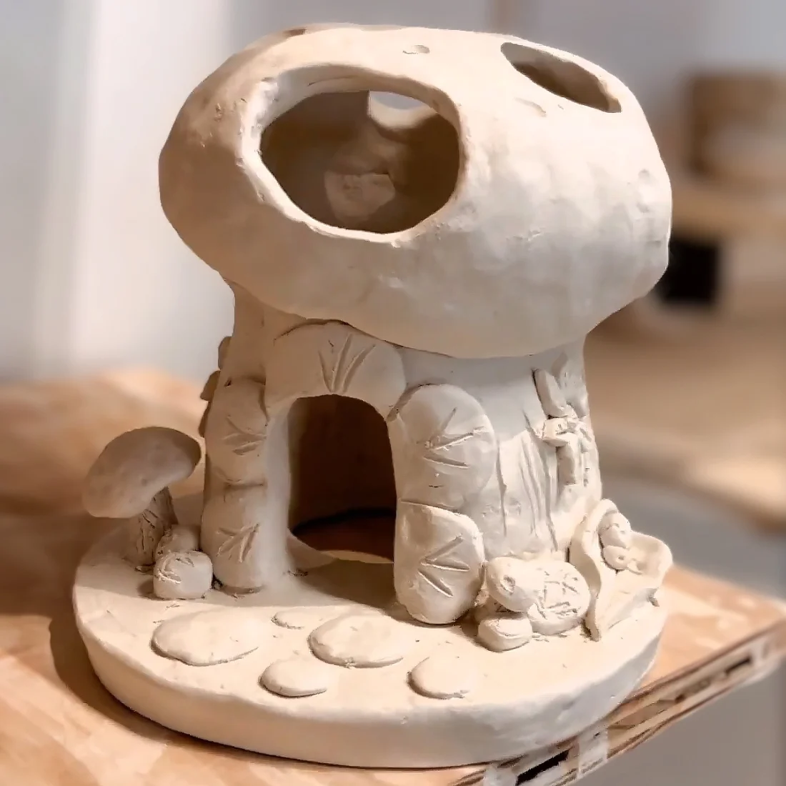 A clay sculpture of a whimsical mushroom house with a wide door, round windows, stone details, and small mushrooms and plants on a circular base, set on a wooden surface.