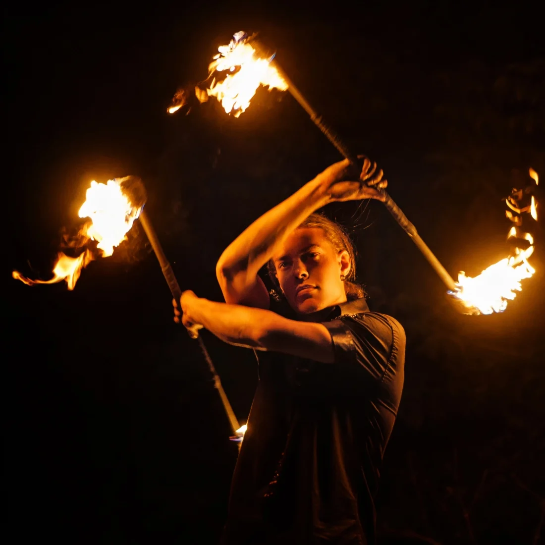 A person performs a fire dance at night, skillfully twirling two flaming batons, creating dynamic patterns of light against a dark background.