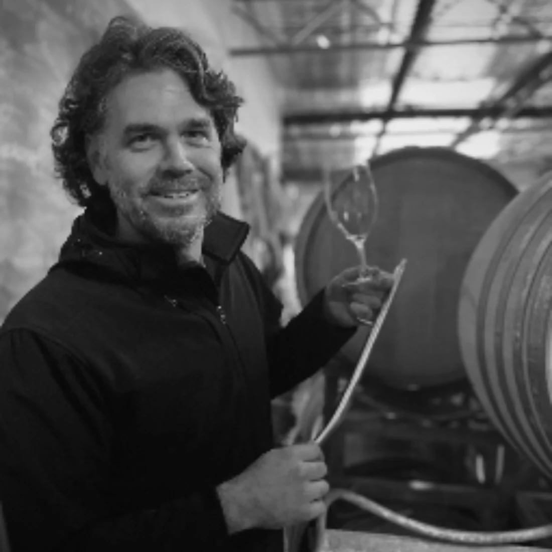 A man with curly hair and a beard smiles while holding a wine glass and a siphon tube in a winery, standing next to large wooden barrels. The image is in black and white.