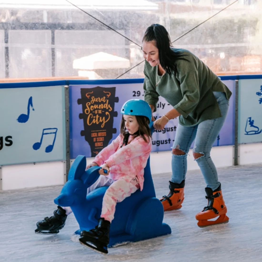 A woman wearing ice skates pushes a young girl, also in skates and a blue helmet, who is sitting on a blue kangaroo-shaped skating aid at an indoor ice rink. Both appear to be enjoying the activity.