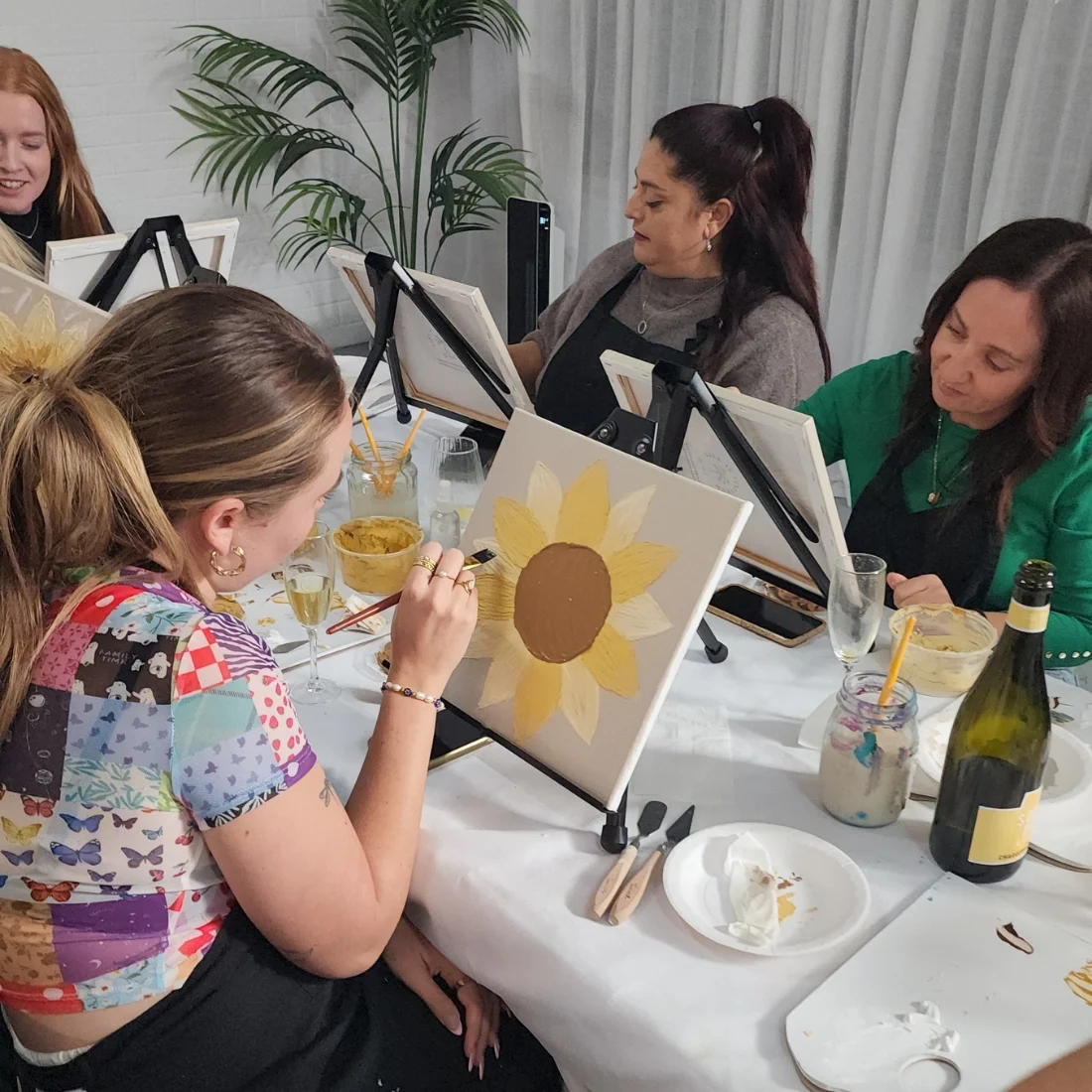Four women sit around a table at a painting class, each working on a canvas with a sunflower design. Art supplies, drinks, and a bottle of wine are on the table. A plant and curtains are in the background.