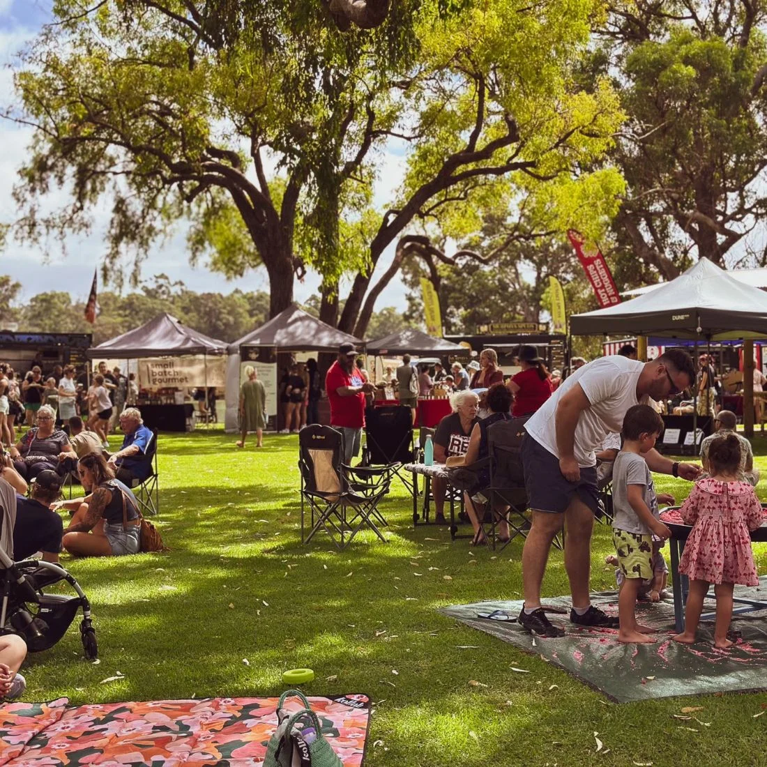 People gather at an outdoor market, sitting on blankets and chairs on the grass. Food stalls and tents are set up under large trees, with families and children enjoying various activities.