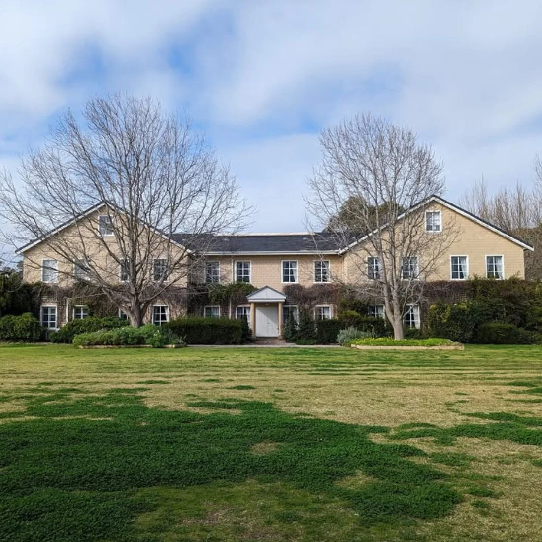 A large, two-story beige house with a white door and windows, surrounded by trees and bushes, stands behind a spacious green lawn under a partly cloudy sky.