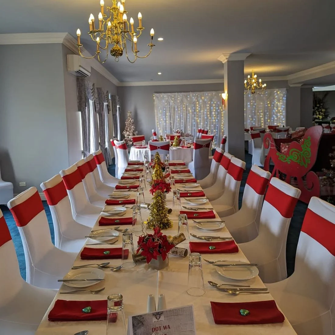 A long dining table set for a festive event, decorated with red napkins, gold and red centerpieces, and white chair covers with red sashes. Chandeliers, holiday decor, and twinkling string lights add a warm ambiance.