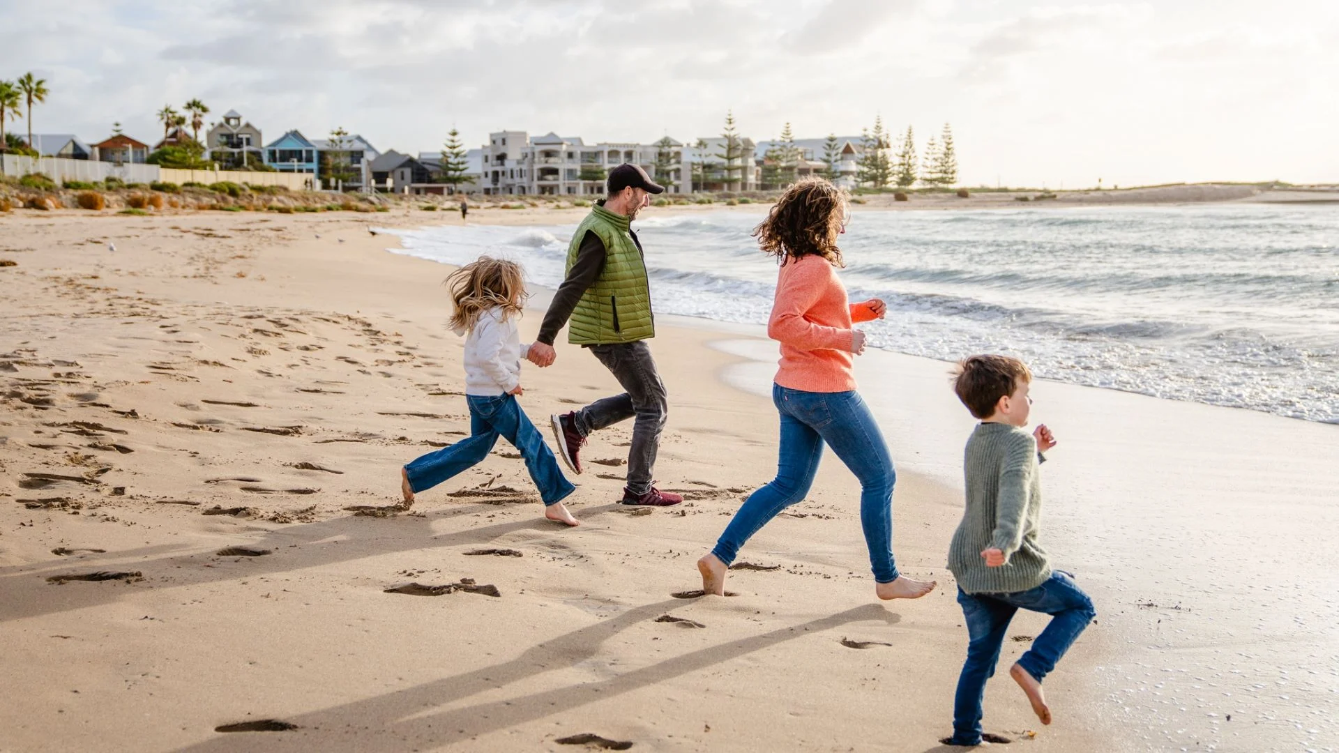 A man, woman, and two children run barefoot along a sandy beach near the ocean, with waves approaching and buildings visible in the background.