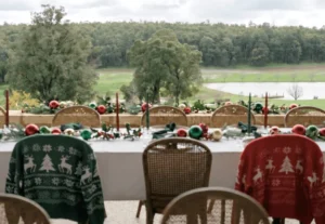 A decorated outdoor table with Christmas-themed sweaters draped over chairs overlooks a green landscape with trees and a lake in the background.
