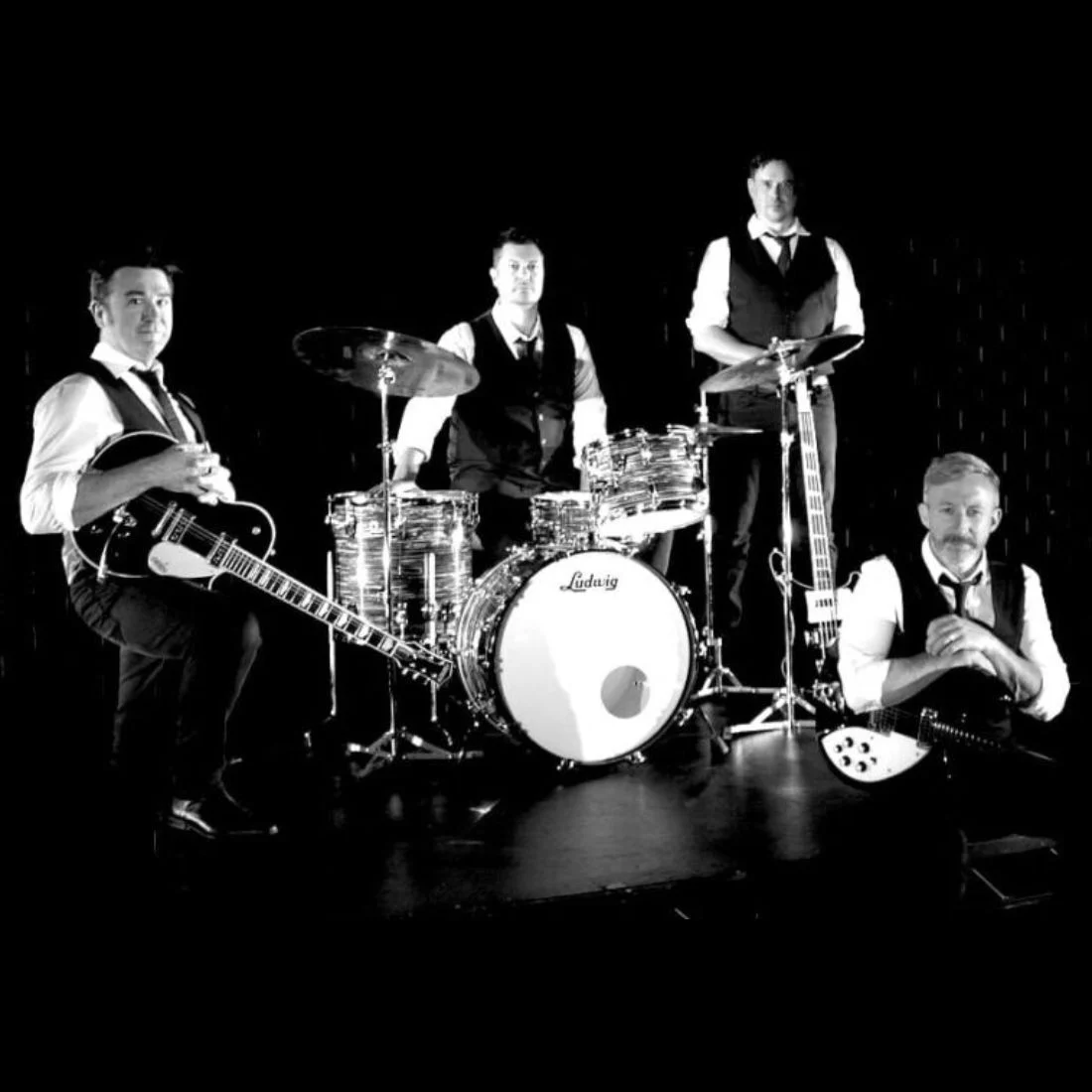 Four men in formal vests and ties pose with musical instruments on a dark stage. One holds a guitar, another sits at a drum set, a third stands with a bass, and the fourth sits on the floor with a guitar.