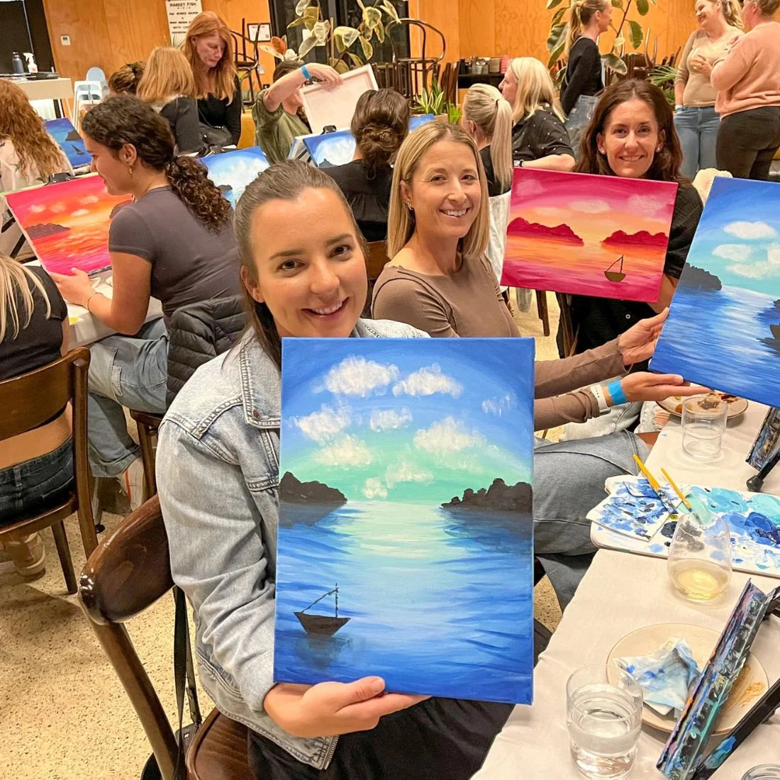 A group of women at a painting class smile and display their colorful seascape paintings with boats, mountains, and sunsets while sitting at tables with art supplies in a lively, indoor setting.