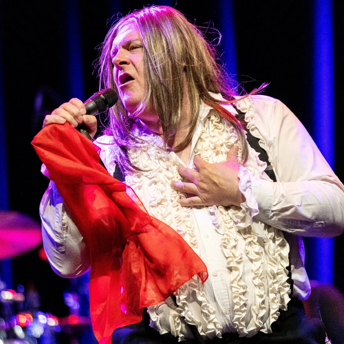 A performer with long hair, wearing a ruffled white shirt and holding a red scarf, passionately sings into a microphone on stage under bright lights.