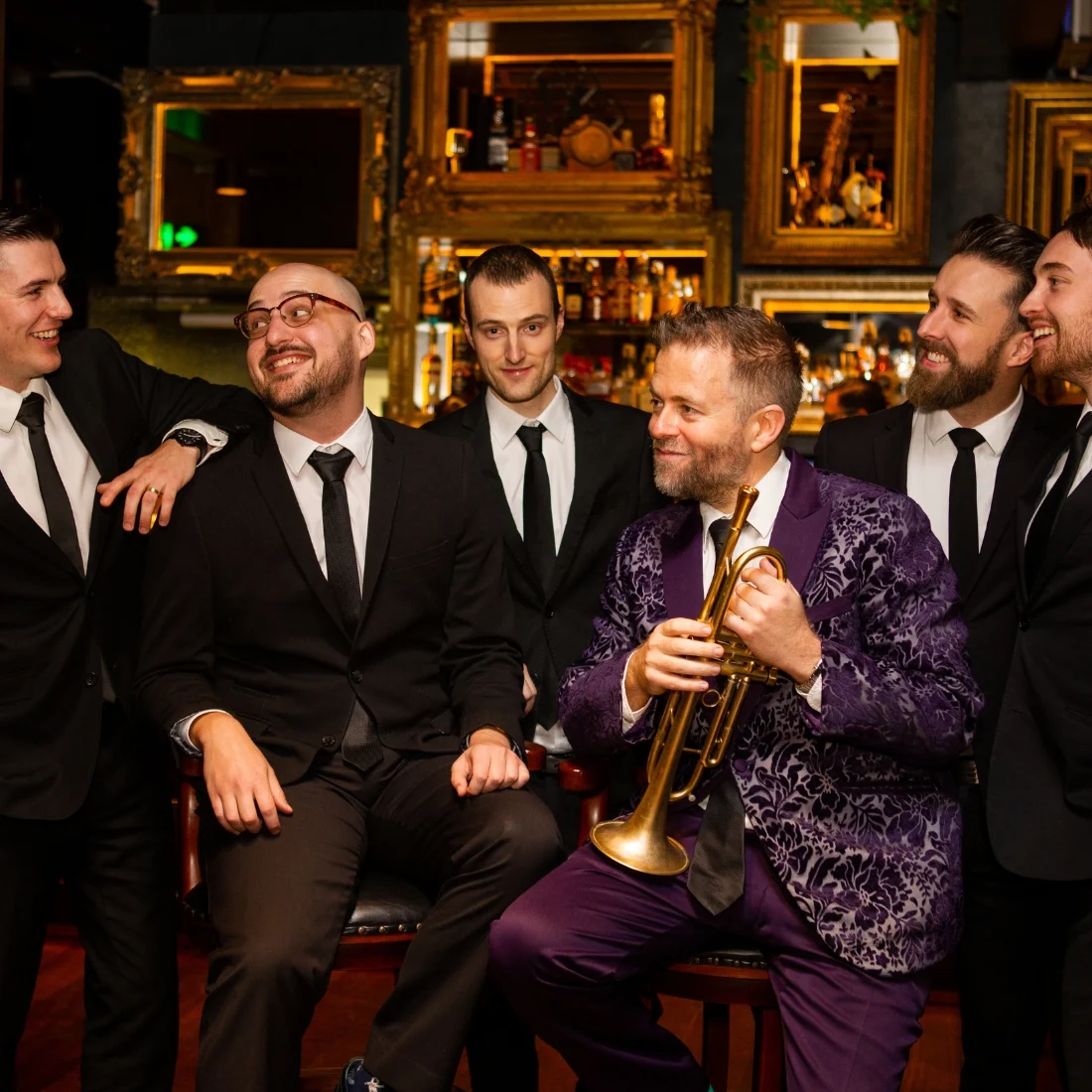 Six men dressed formally are laughing and smiling together indoors; five wear black suits and ties, while one in a purple velvet suit with a trumpet sits at the center. They appear to be enjoying a lively, friendly moment.